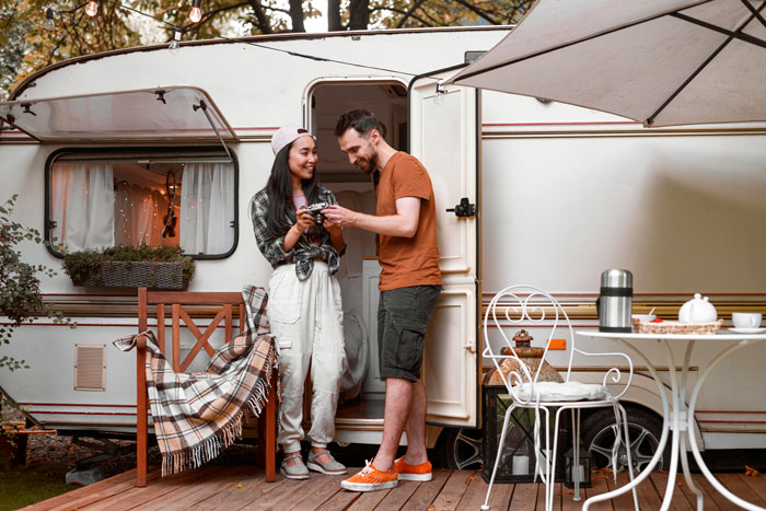 Couple smiling outside a camper, with a table set for coffee; surrounded by trees and nature.