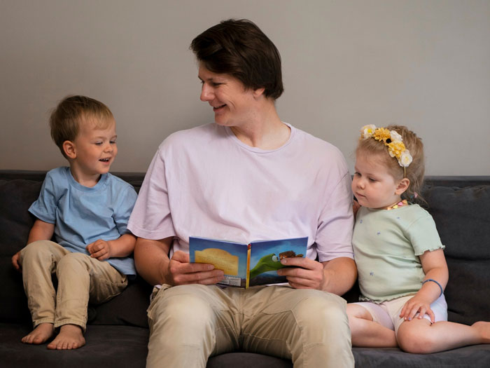 Man babysitting sister’s kids on couch, reading a book, illustrating man agrees to babysit sister’s kids trans narrative.