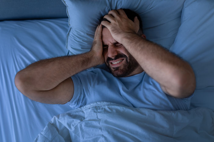 Man in bed holding his head in discomfort, disturbed by noisy coffee grinder causing neighbor complaint at night.