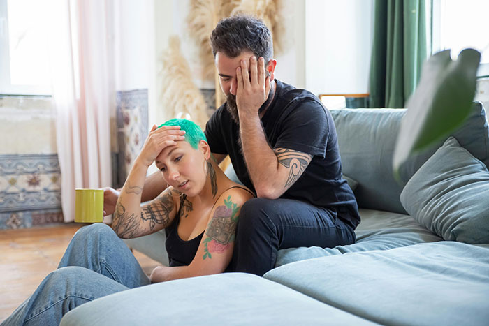 Distraught couple on sofa after altercation about neglectful MIL and CPS threat, with woman holding a mug.