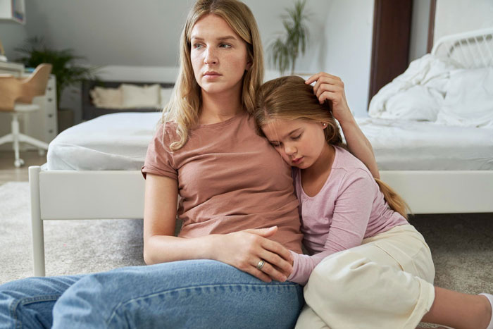Worried mother comforting 7-year-old girl experiencing lactose intolerance symptoms at home.